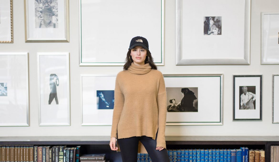 Creativity Takes Courage - woman in a hat and yellow sweater standing in front of a wall of art and books