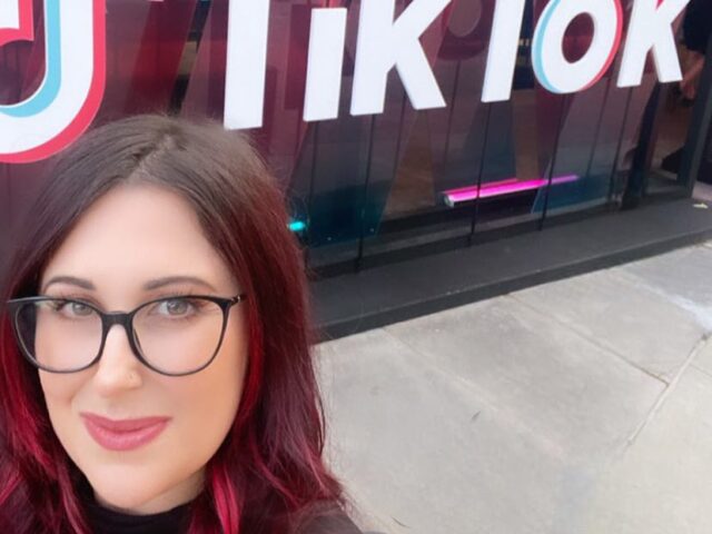 Jennifer Paukman stands in front of TikTok sign. She has red hair and wears glasses.